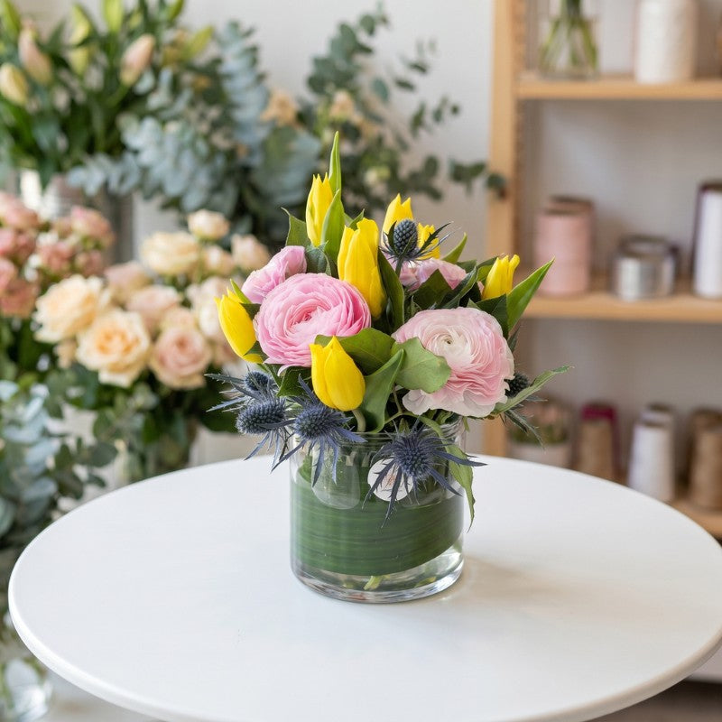 Yellow tulip and pink ranunculus floral arrangement with blue thistle accents in glass vase