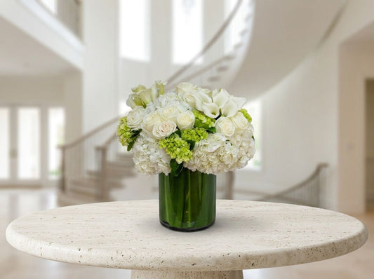 Luxurious white floral arrangement featuring ivory roses, white calla lilies, lush hydrangeas, and fresh green accents in a dark green glass cylinder vase, displayed on an elegant marble table.