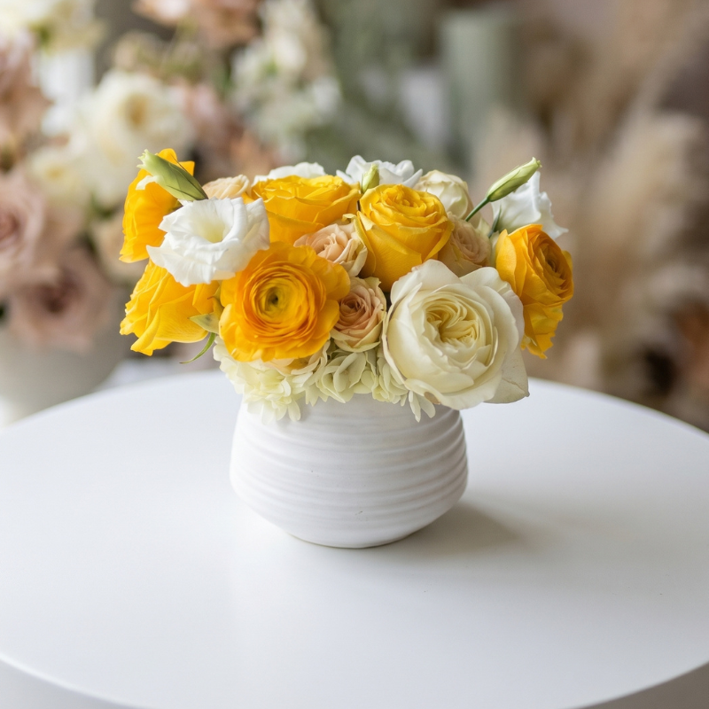 Compact floral arrangement featuring vibrant yellow roses and ranunculus, ivory garden roses, cream spray roses, and white lisianthus in a modern white ceramic vase.