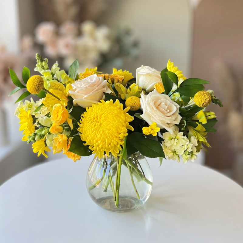 Cheerful yellow and white floral arrangement featuring yellow chrysanthemums, cream roses, yellow daisies, craspedia, snapdragons, and seasonal greenery in a clear glass vase.