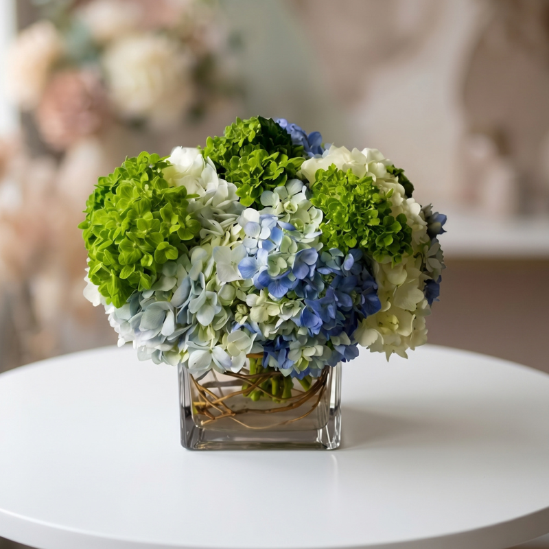 Elegant newborn flower arrangement featuring blue and white hydrangeas with green hydrangea accents in a modern glass cube vase, perfect for welcoming a new baby boy.