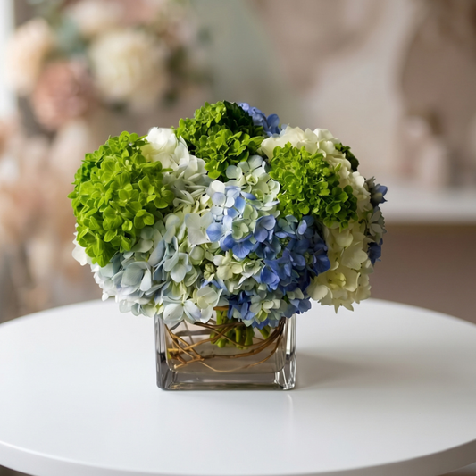 Elegant newborn flower arrangement featuring blue and white hydrangeas with green hydrangea accents in a modern glass cube vase, perfect for welcoming a new baby boy.
