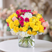Vibrant floral arrangement featuring yellow roses, hot pink spray roses, blush peach roses, and white hydrangeas in a clear glass cylinder vase, designed for birthdays, celebrations, and cheerful home décor