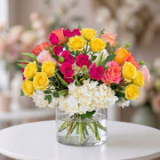 Vibrant floral arrangement featuring yellow roses, hot pink spray roses, blush peach roses, and white hydrangeas in a clear glass cylinder vase, designed for birthdays, celebrations, and cheerful home décor