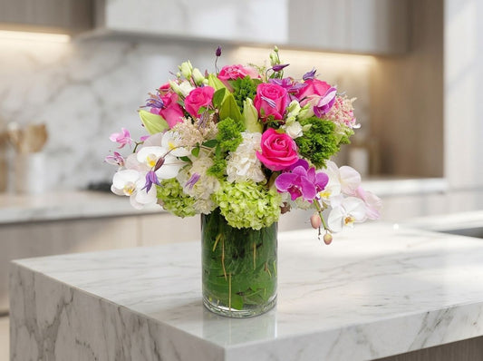 Luxurious pink and white floral arrangement featuring vibrant pink roses, cascading white and purple orchids, lush white and green hydrangeas, delicate purple accents, and fresh greenery in an elegant clear glass cylinder vase.