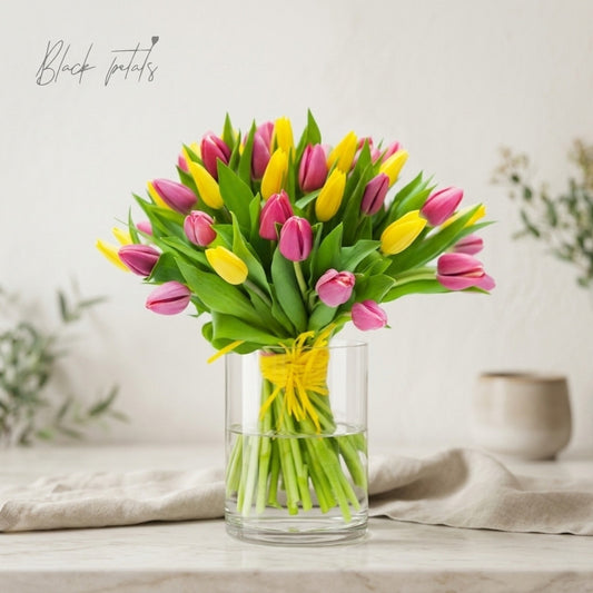 Pink and yellow tulip bouquet arranged in a clear glass vase with green foliage, displayed on a neutral table setting for a spring floral arrangement.