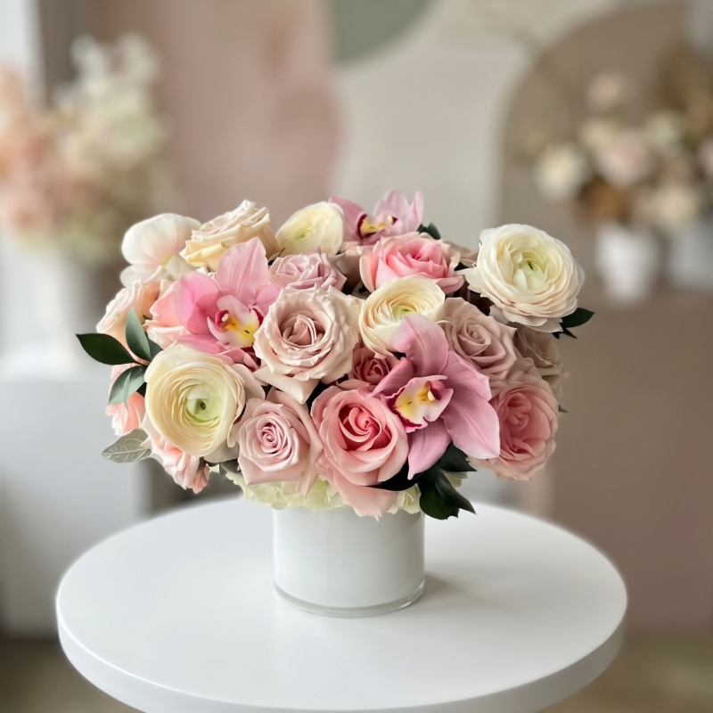 Luxury newborn flower arrangement featuring blush pink roses, white ranunculus, and pink cymbidium orchids in a modern white vase, designed to celebrate the arrival of a new baby.