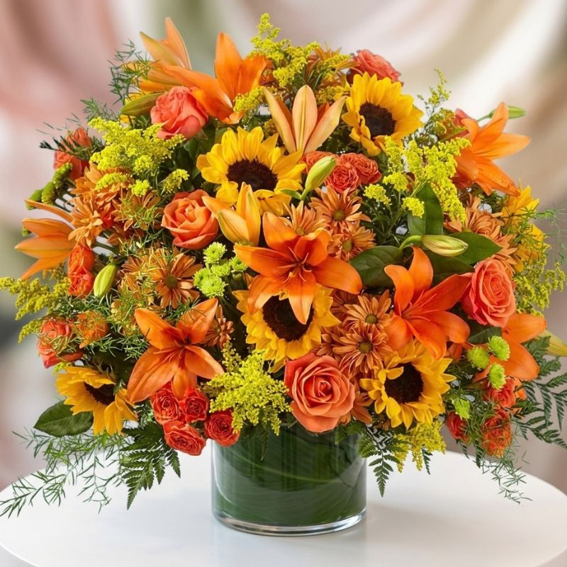 Fall flower arrangement with orange lilies, sunflowers, coral roses, bronze chrysanthemums, yellow filler flowers, and greenery in a leaf-lined glass vase.