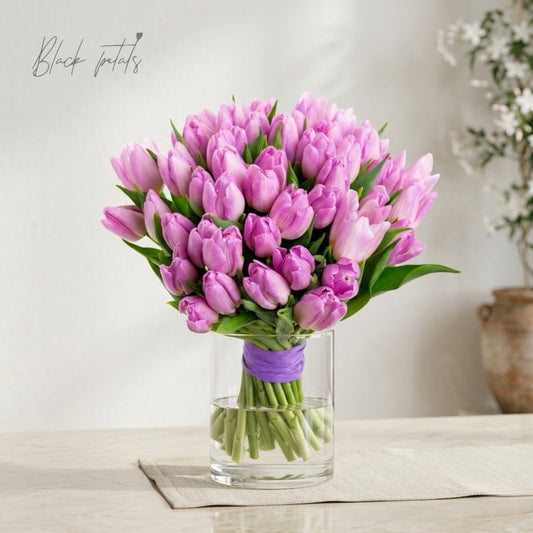 Pink tulip bouquet arranged in a clear glass vase with a purple wrap, displayed on a neutral table for an elegant spring flower arrangement.