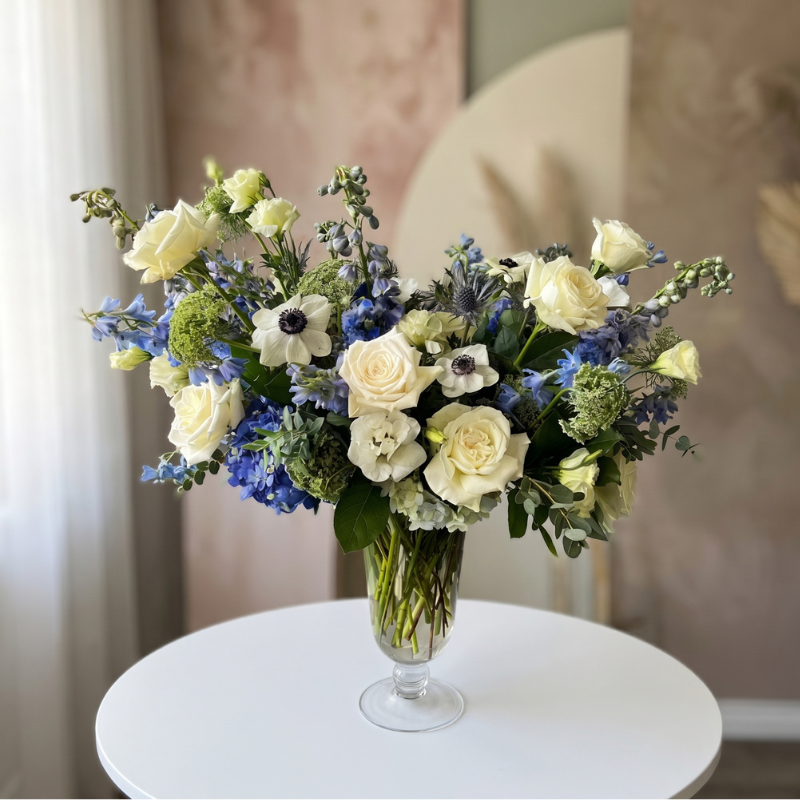 Elegant newborn flower arrangement featuring white roses, blue delphinium, white anemones, lisianthus, and seasonal blooms in a clear glass vase, perfect for celebrating a new baby boy.