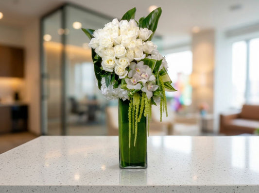 Elegant white floral arrangement featuring lush ivory roses, delicate white orchids, white hydrangeas, cascading green amaranthus, and glossy tropical greenery in a tall rectangular green glass vase.