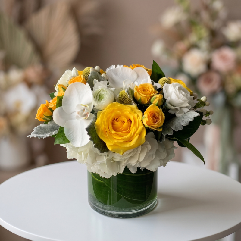 Cheerful newborn flower arrangement featuring yellow roses, white orchids, white hydrangeas, ranunculus, and seasonal blooms in a modern glass vase, perfect for celebrating a new baby.