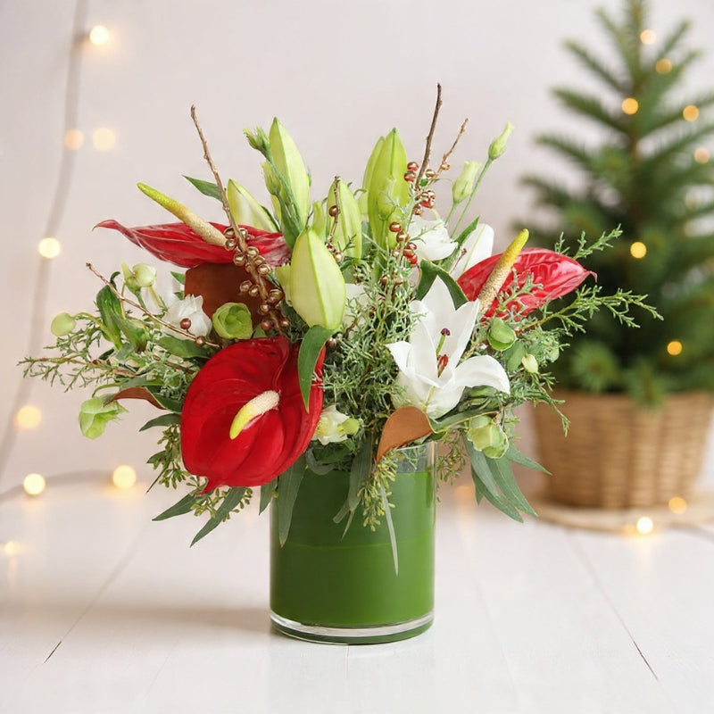 White lilies, red anthuriums, roses, and winter greenery arranged in a tall vase for a festive Christmas bouquet.