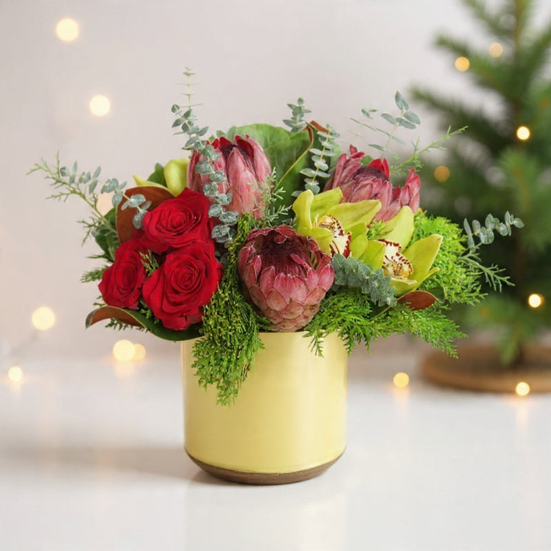 Protea blooms with roses, orchids, berries, and winter greenery arranged in a cream vase for a modern holiday floral design.