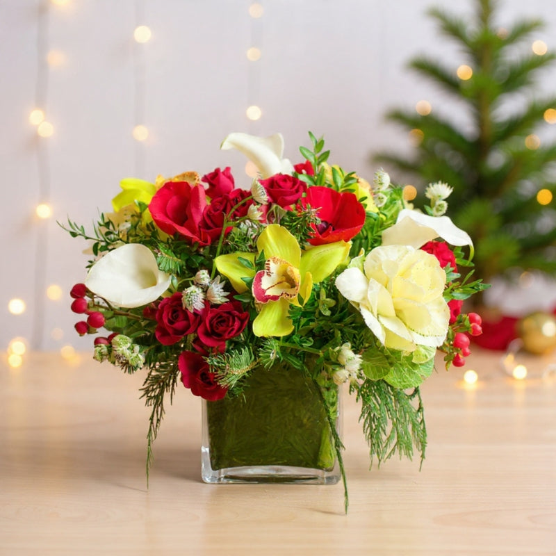 Yellow orchids, red roses, white blooms, pinecones, and winter greenery arranged in a tall vase for bright holiday décor.