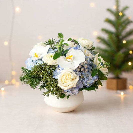 Blue hydrangeas, white orchids, and ivory roses arranged in a clear vase for an elegant winter bouquet.