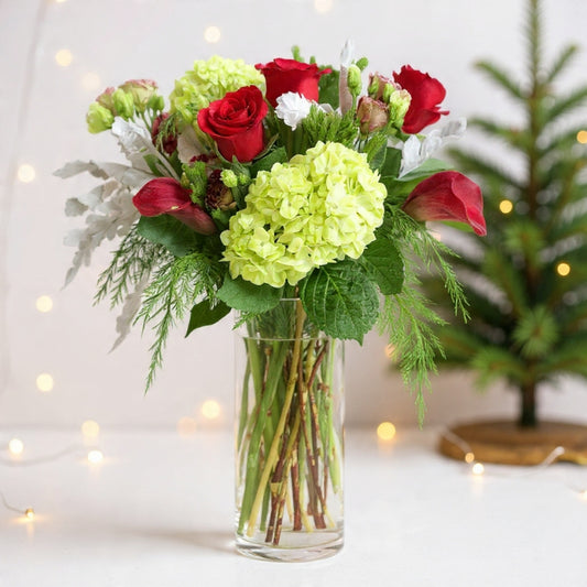 Lime hydrangeas, red roses, and lush winter greenery arranged in a tall vase for a cheerful bright holiday floral arrangement.