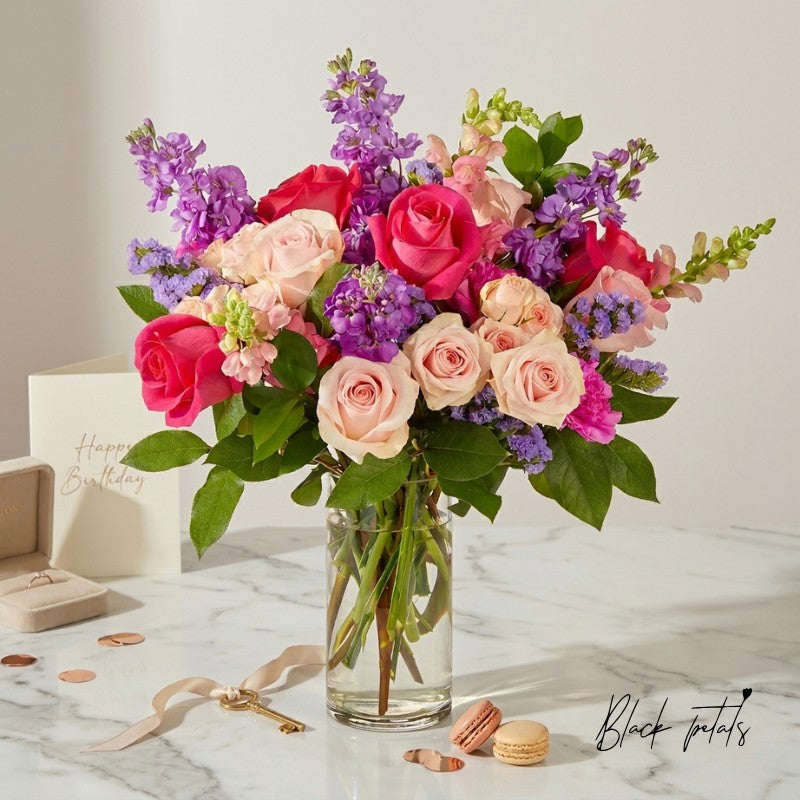 Elegant pink and blush rose bouquet with purple florals arranged in a glass vase, styled on a marble surface with birthday card and luxury accents.