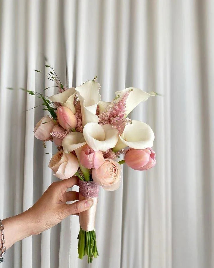 Hand holding elegant bridal bouquet with white calla lilies, pink tulips, and roses against white curtain