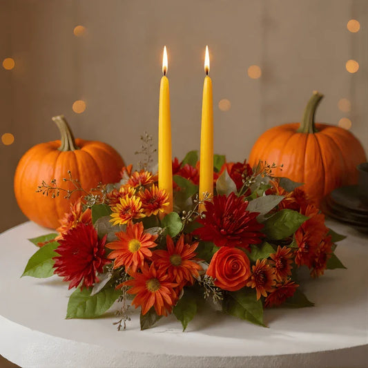 Autumn centerpiece with orange and red flowers, green leaves, two lit yellow candles, and pumpkins on white table
