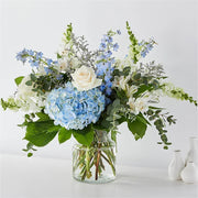 Elegant bouquet with blue hydrangeas, white roses, delphiniums, and greenery in clear glass vase