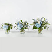 Three clear glass vases with blue hydrangeas and white flowers arranged on a white surface