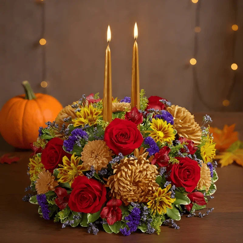 Autumn floral centerpiece with red roses, yellow and orange flowers, purple accents, two lit gold candles, and a pumpkin backdrop