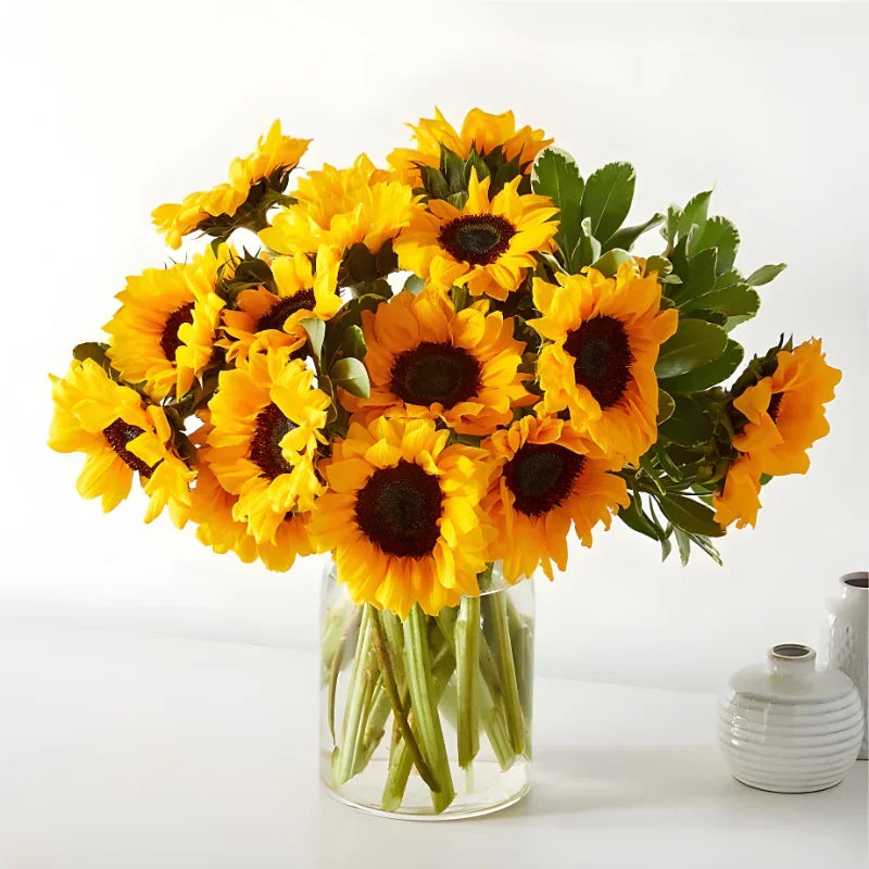 Bright yellow sunflower bouquet with green leaves in a clear glass vase on white table