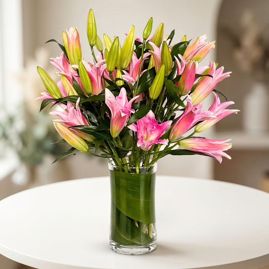 Elegant arrangement of fresh pink lilies with blooming flowers and green buds, complemented by lush greenery in a clear glass cylinder vase, available at Black Petals.