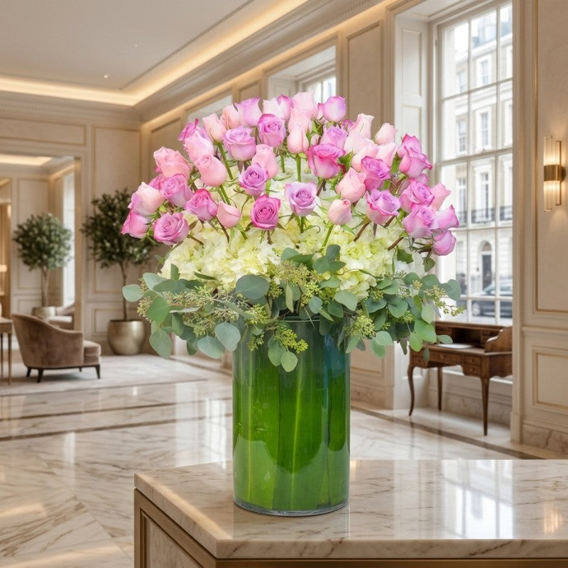Elegant blush pink rose and hydrangea lobby arrangement in a tall green glass vase displayed in a luxury hotel lobby.