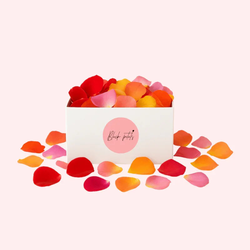 White box filled with multicolor rose petals in red, orange, and pink shades, scattered on a pale pink background