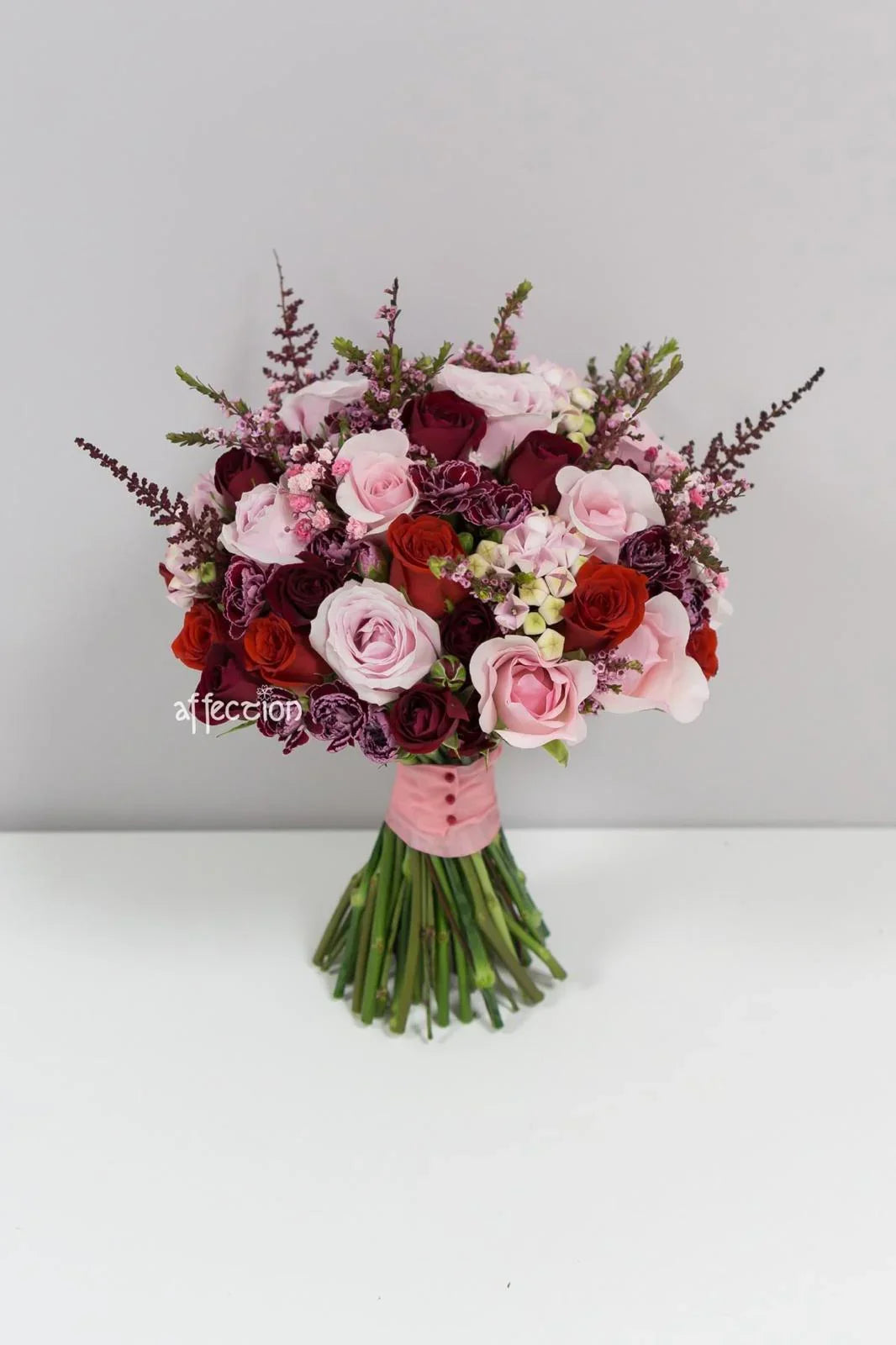 Bridal bouquet with red and pink roses, purple flowers, and green stems wrapped in pink ribbon