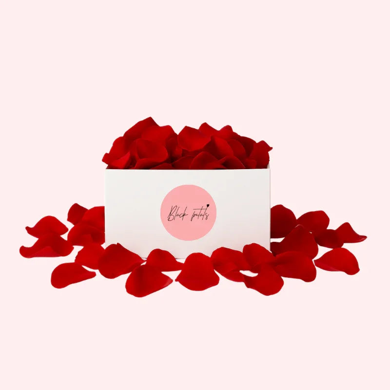 Box filled with vibrant red rose petals and scattered petals around on light pink background