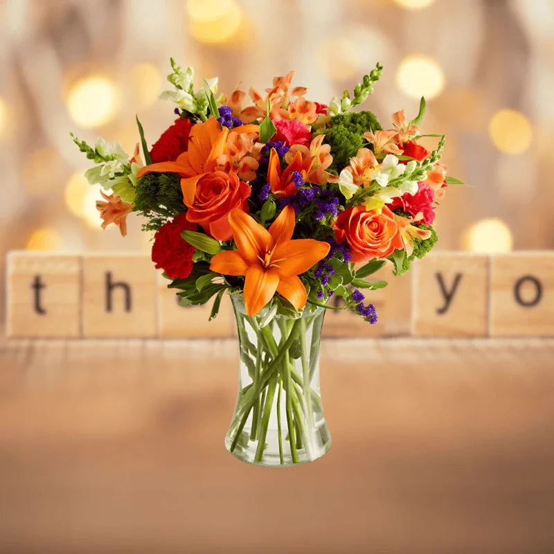 Colorful fall bouquet with orange lilies, roses, and mixed flowers in clear vase on wooden table