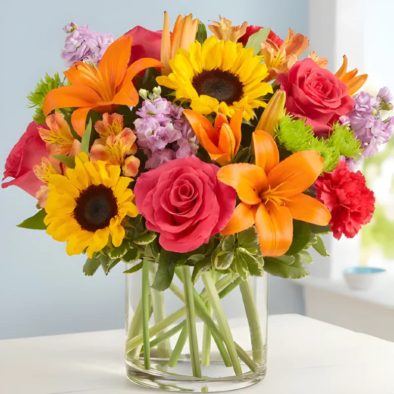 Bright autumn flower arrangement with sunflowers, orange lilies, red roses, and pink carnations in clear glass vase