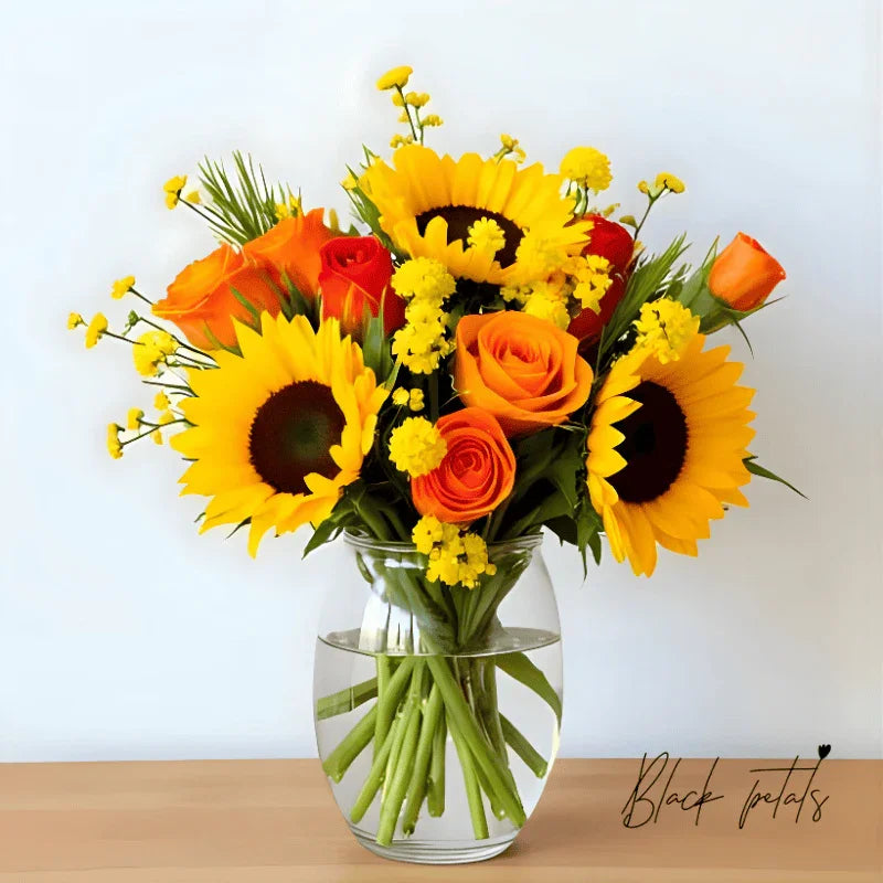 Bright bouquet of sunflowers, orange roses, and yellow filler flowers in clear glass vase on wooden surface