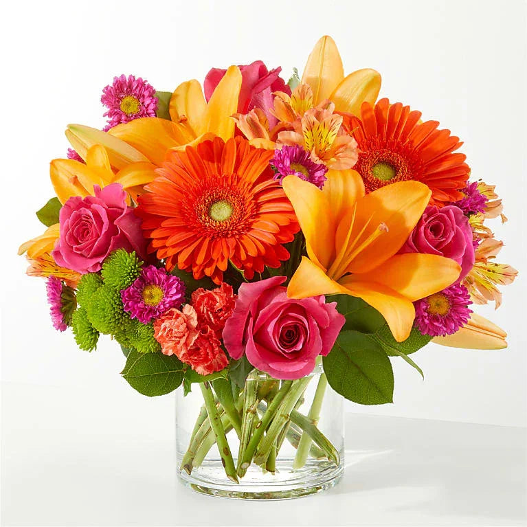 Bright mixed flower bouquet with orange lilies, gerbera daisies, pink roses, and green accents in a clear glass vase