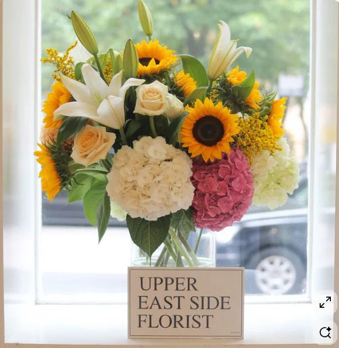 Bright bouquet of sunflowers, lilies, roses, and hydrangeas in clear vase with florist sign on windowsill