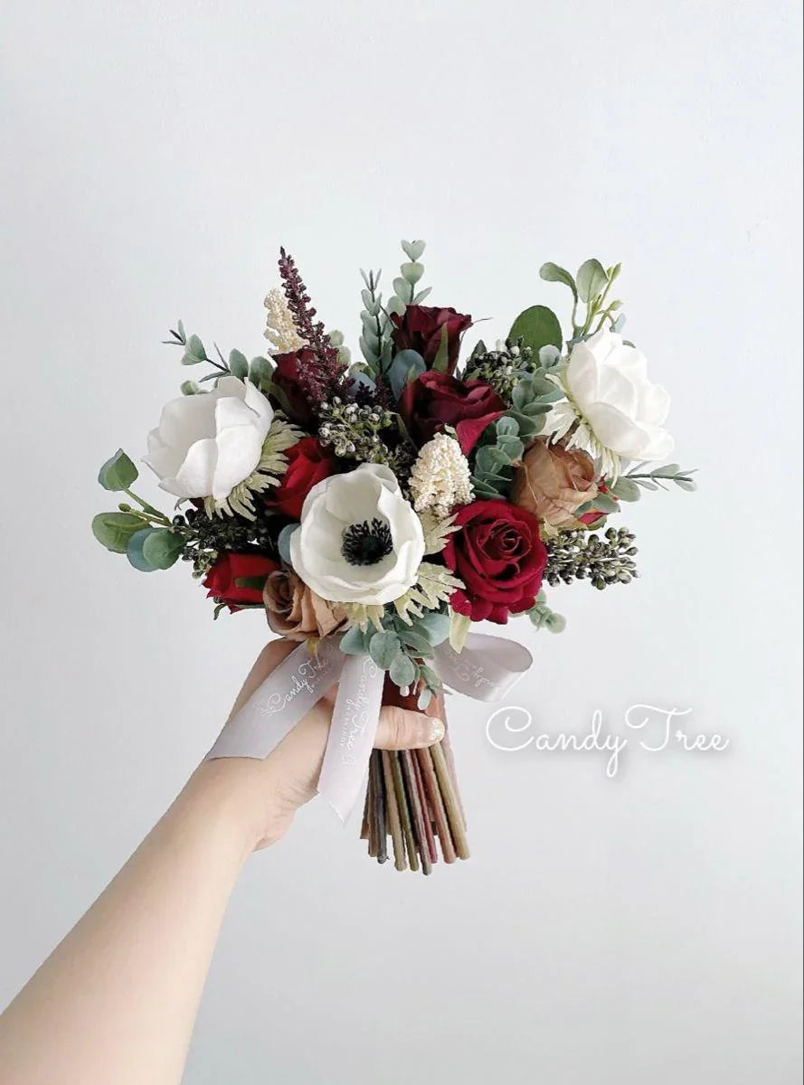 Hand holding a burgundy and white bridal bouquet with roses, anemones, and greenery on white background